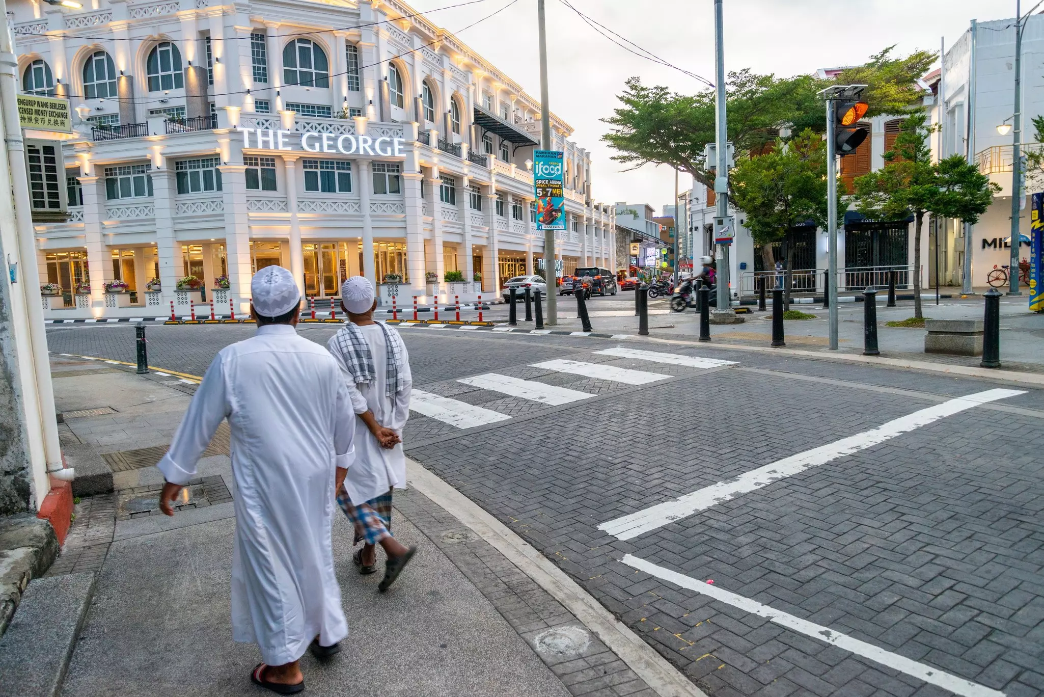 Two men in white tunics are pictured from behind walking on a city street toward a historic hotel.