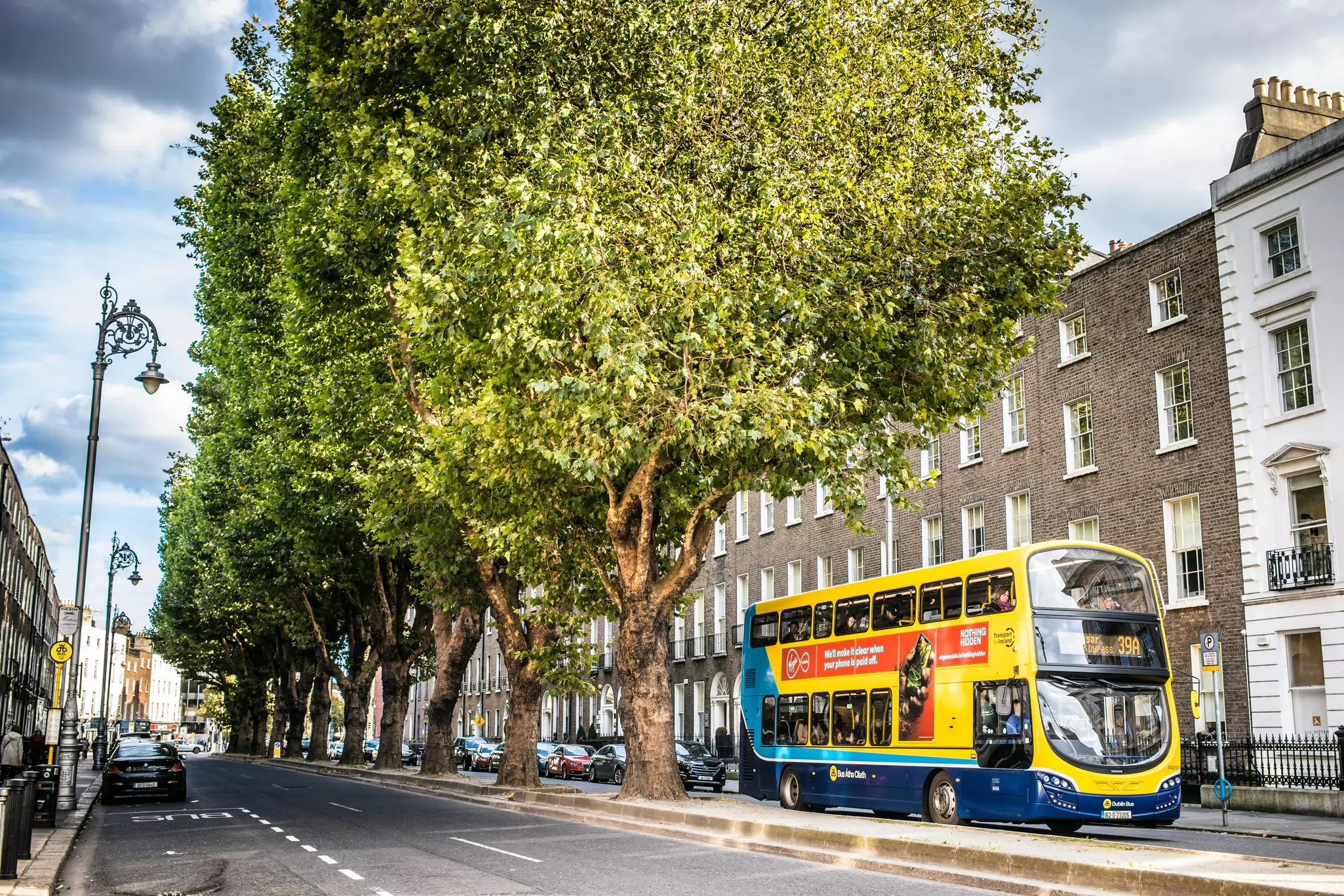 A double-decker bus drives past historic brick buildings on a city street. Tall trees grow in a median in street.