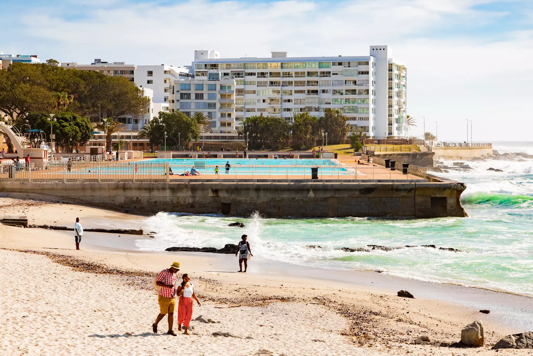 People walk on the sand on a city beach. A public swimming pool stands at the water’s edge, and apartment buildings are seen in the background.