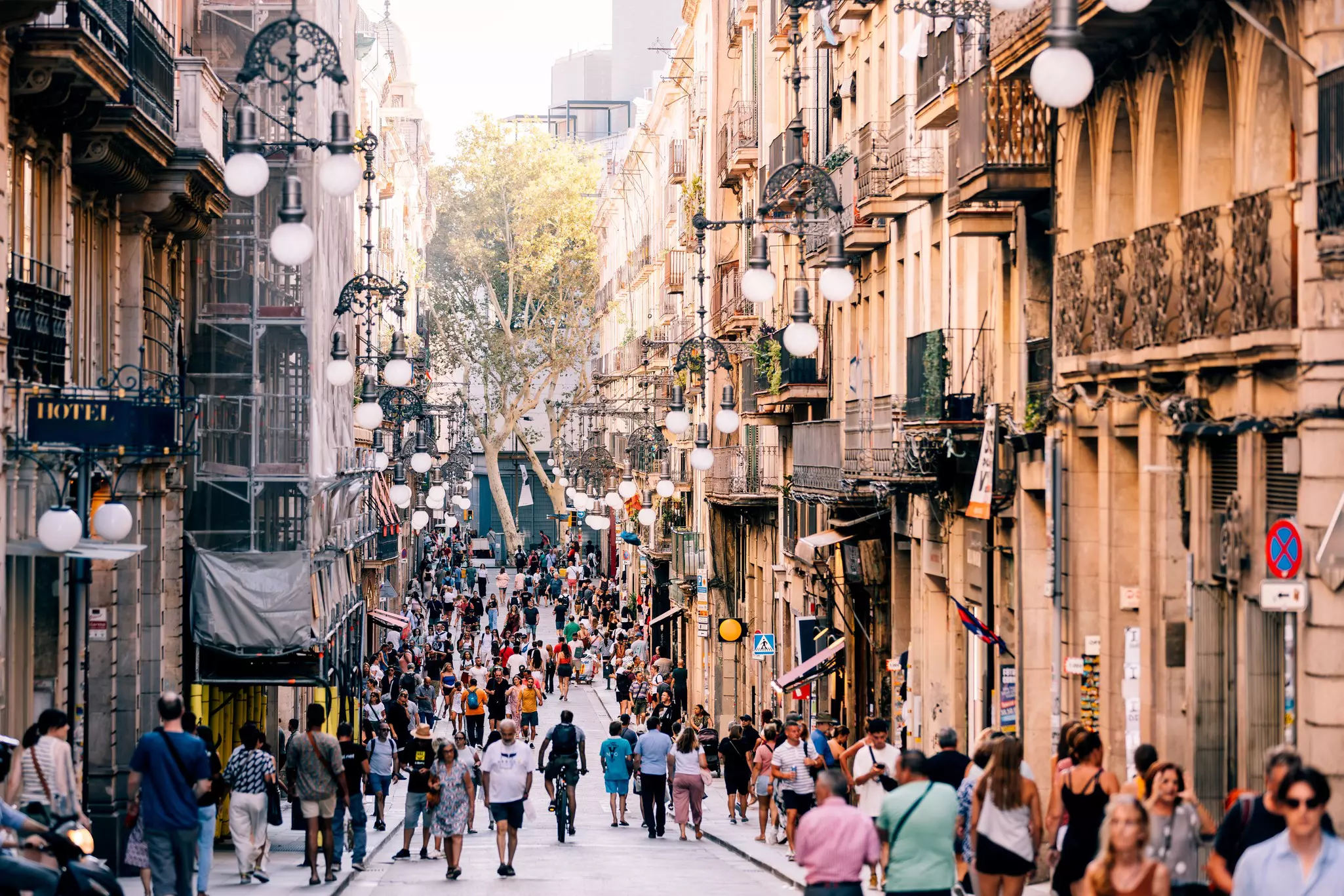People walk down a narrow street in a city, with streetlights and wrought-iron balconies of buildings seen in the view.