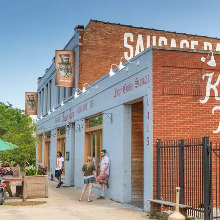 A side view of a brick building, with the storefront painted light gray; people are leaning on the wall.