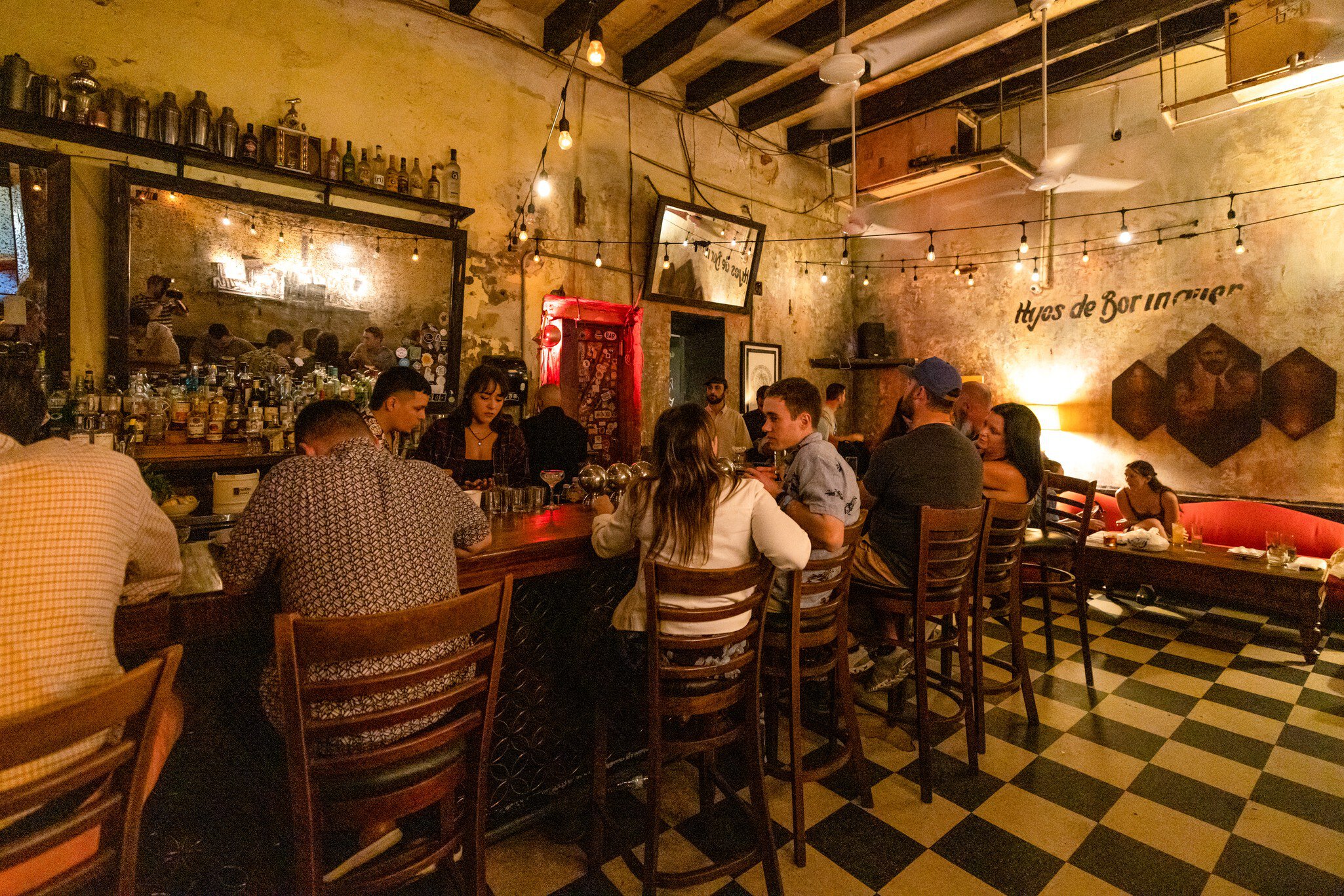 People sit and drink cocktails at at a bar which has a green and white tiled floor and a bar