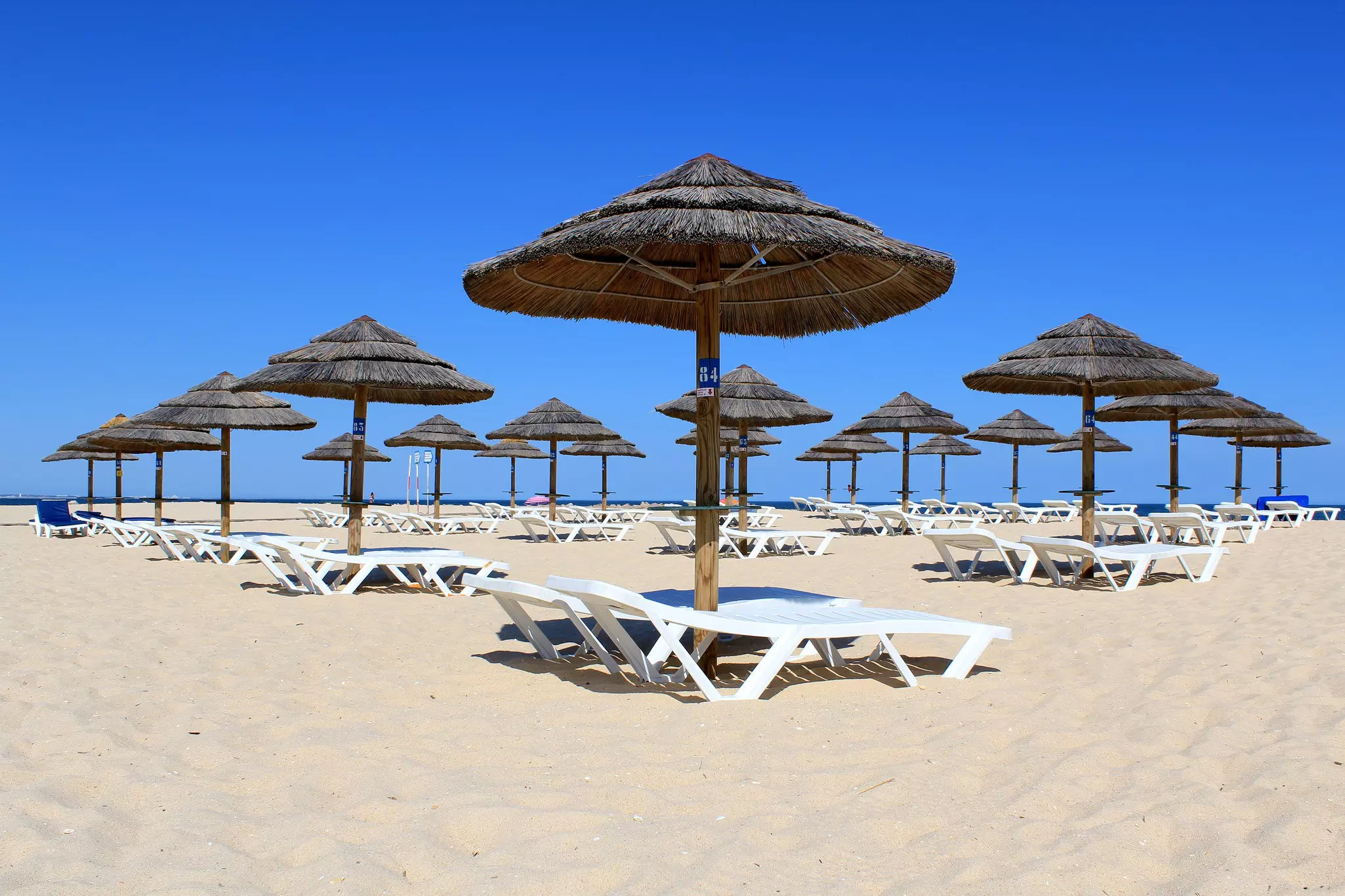 A series of white sun loungers and grass-topped umbrellas on a wide, sandy beach.