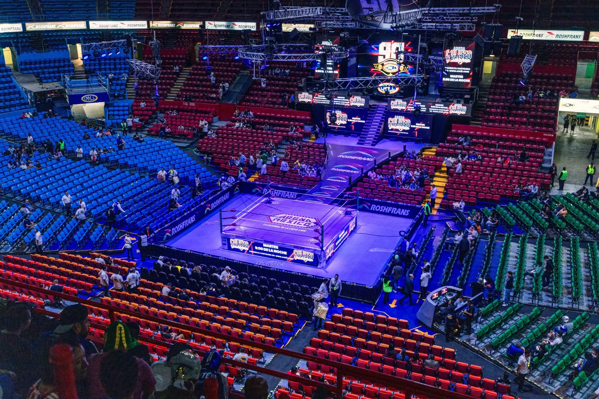 Red, green and blue seats surround a blue mat and wrestling ring in an arena with lots of lights and people coming in.