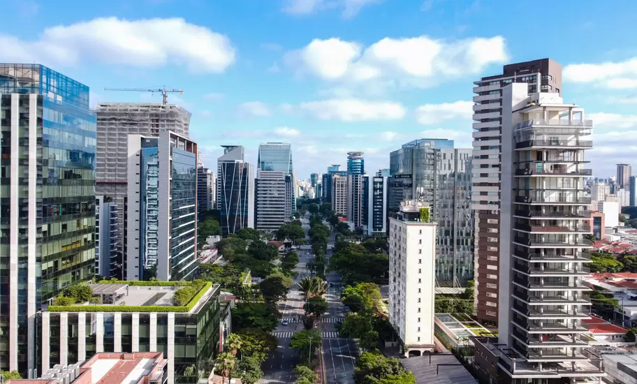 São Paulo high-rise towers lining a tree-filled avenue, with a crane atop a building under construction and blue sky