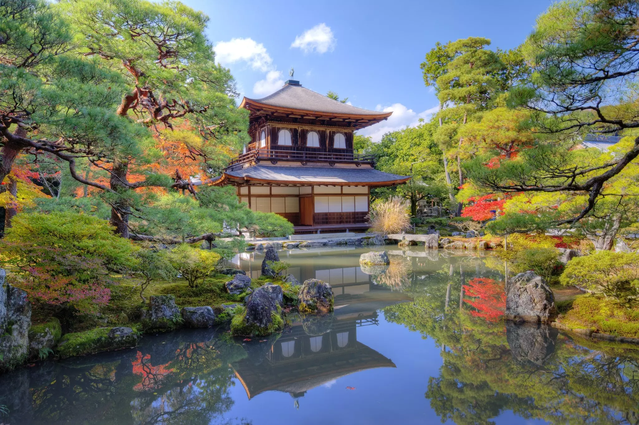 A temple is pictured in front of a placid pond surrounded by trees bearing fall foliage.