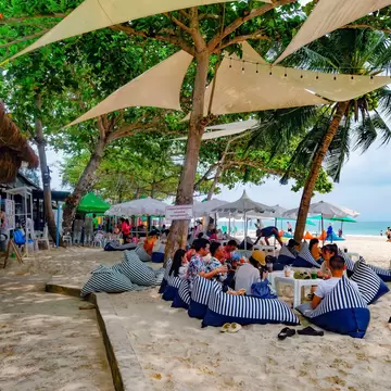 People lounging on striped beanbags under white sail shades and trees on a sandy beach, with the sea and swimmers behind.