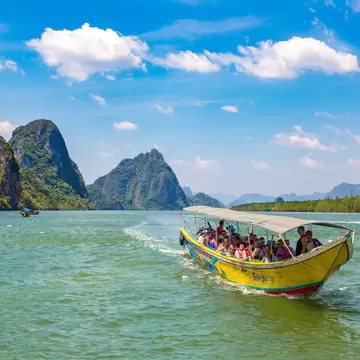 A yellow longtail boat full of passengers crossing green water toward tall limestone islands in Phang-Nga Bay, Thailand.