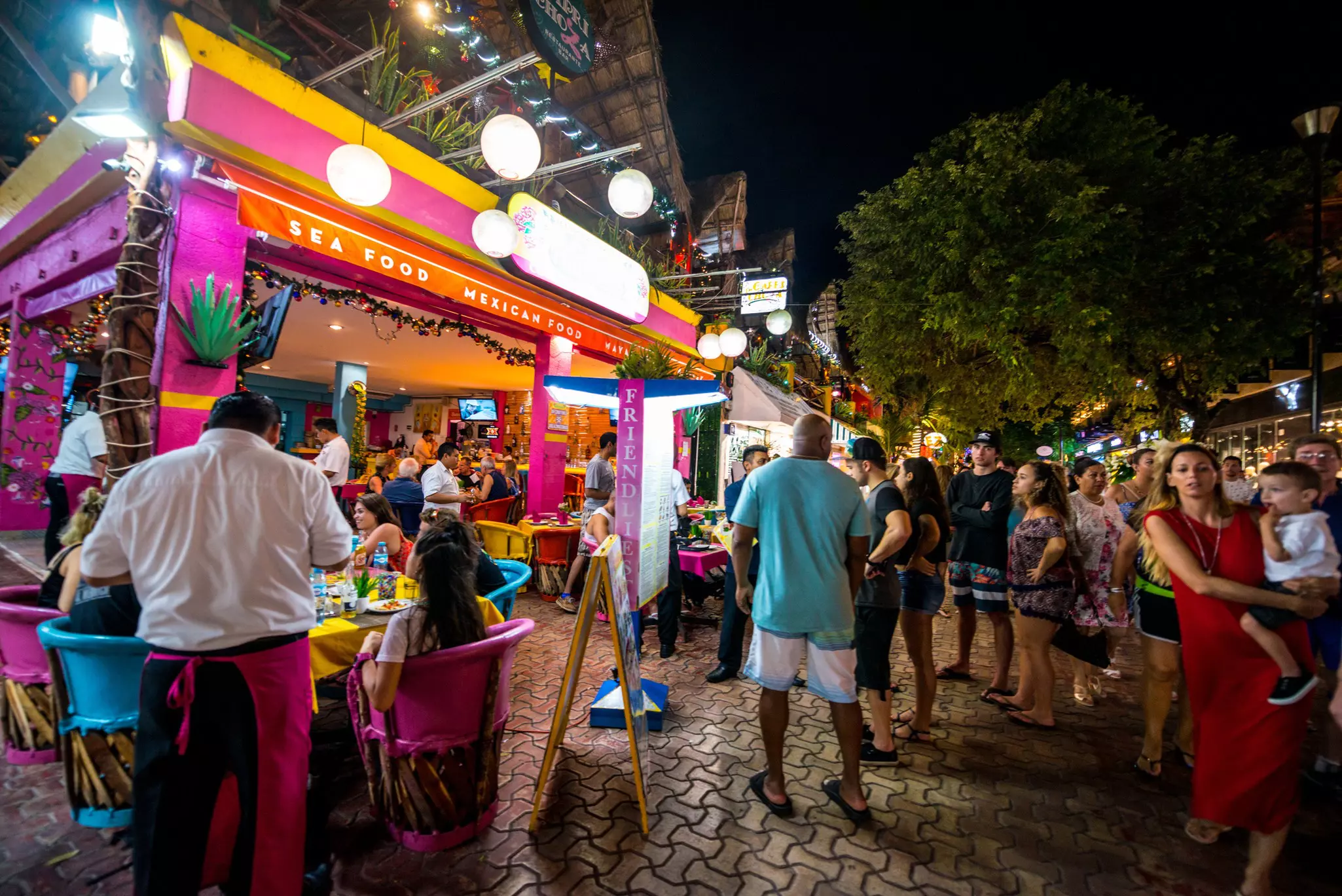 People in a warm-weather place wait on a street outside a restaurant at night.