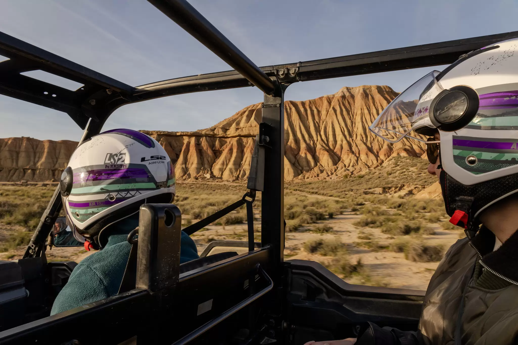 Two riders in helmets sit in a buggy riding through a badlands-desert-like landscape