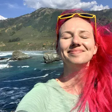 A woman with red hair smiling with her eyes closed with the Big Sur coastline behind her