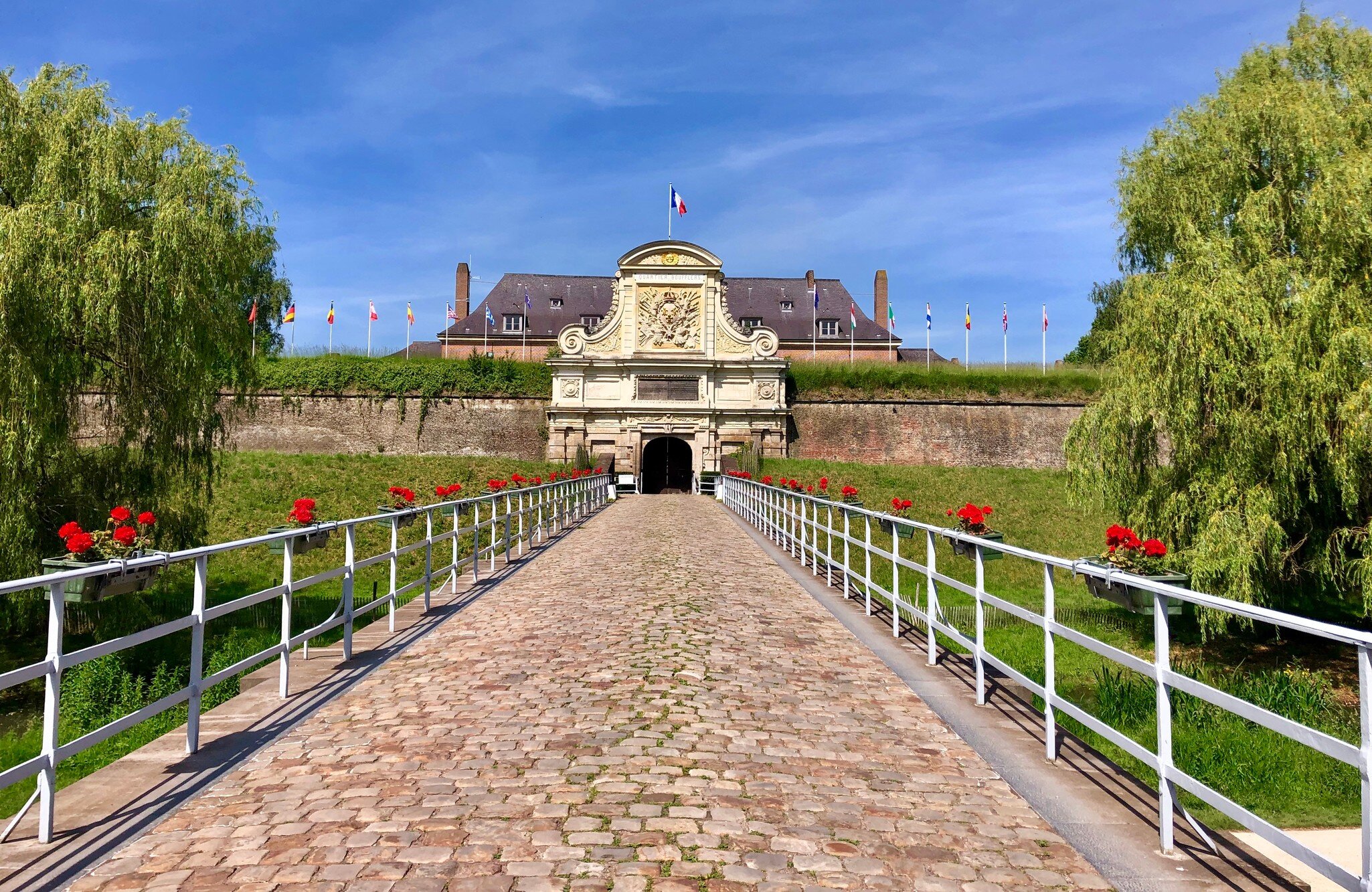 A cobbled causeway lined with white railings and red geraniums leading to an ornate stone gateway set into a grassed rampart, flags flying behind.