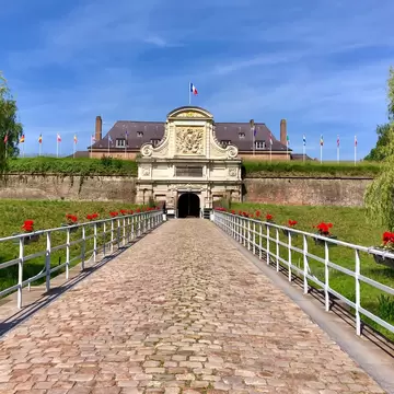 A cobbled causeway lined with white railings and red geraniums leading to an ornate stone gateway set into a grassed rampart, flags flying behind.