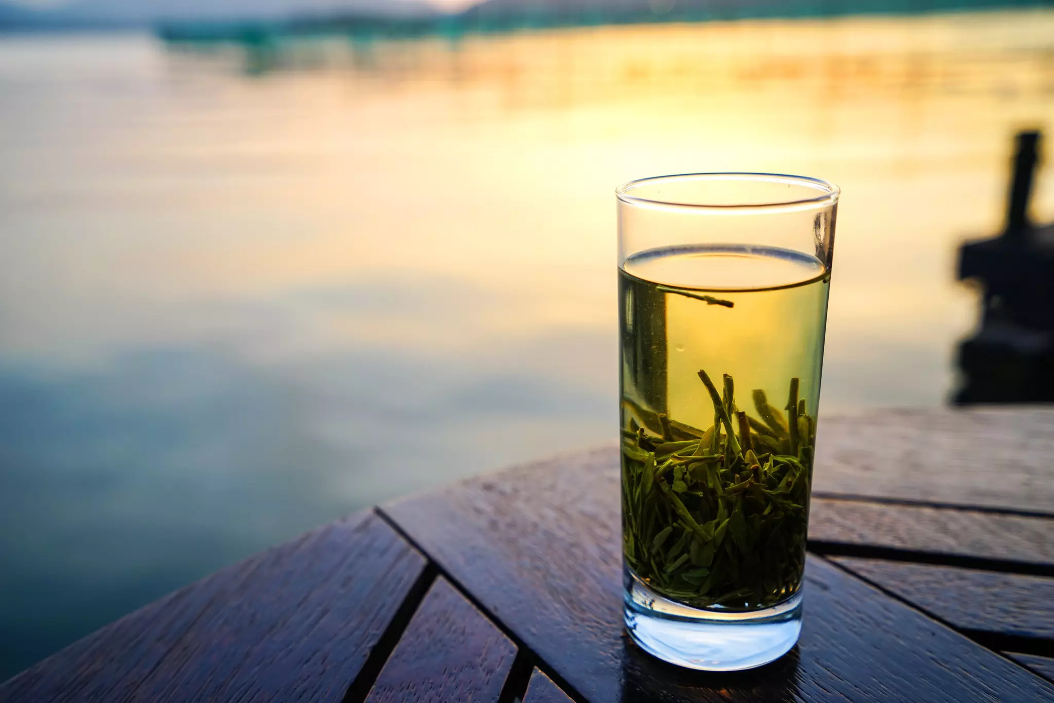 Close-up of a glass of green Longjing tea on a wooden table with the West Lake in the background.