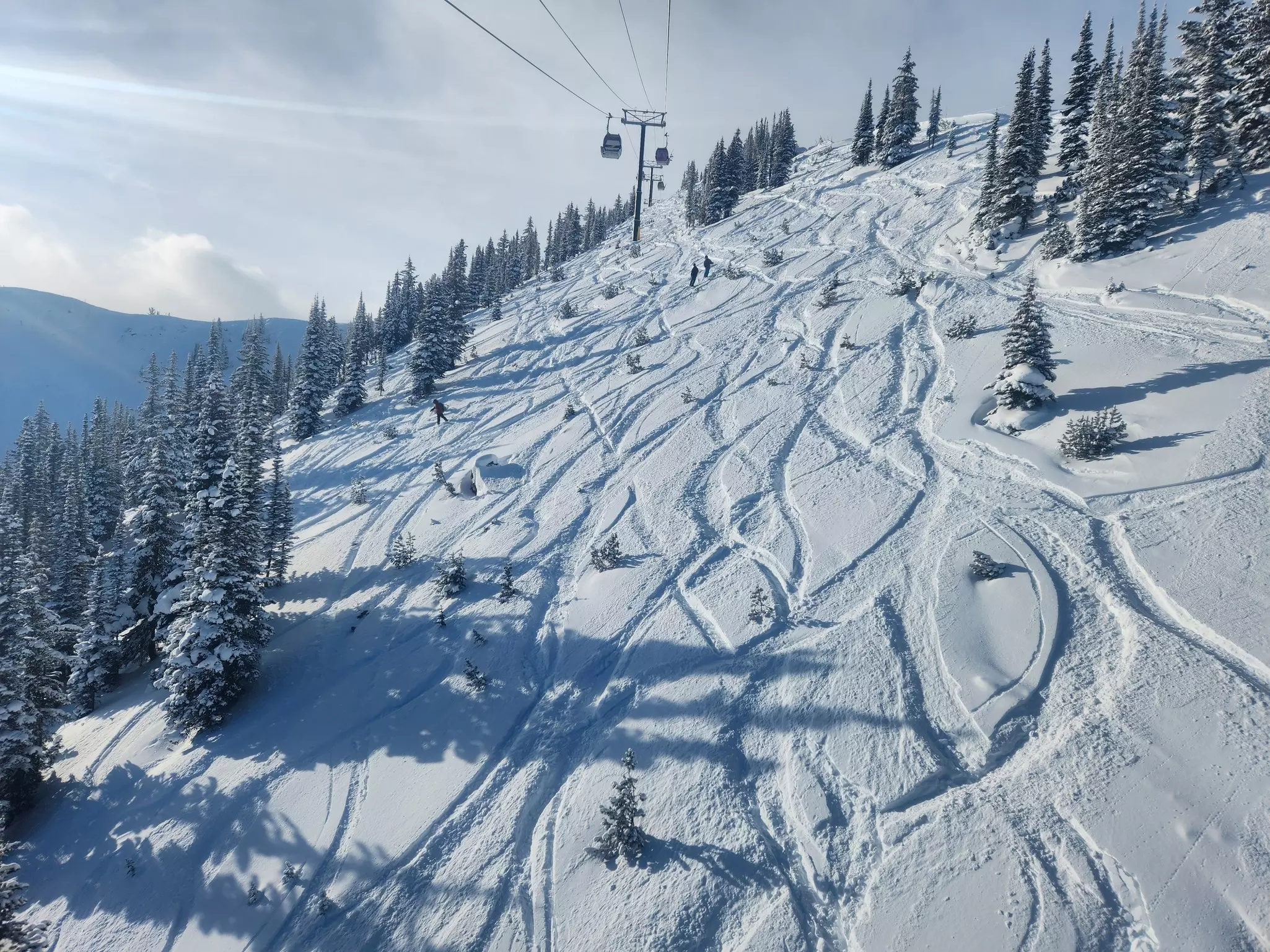 Aerial view of ski slope with tracks going down the mountain, evergreens covered in snow and a ski lift above on a mostly sunny day.