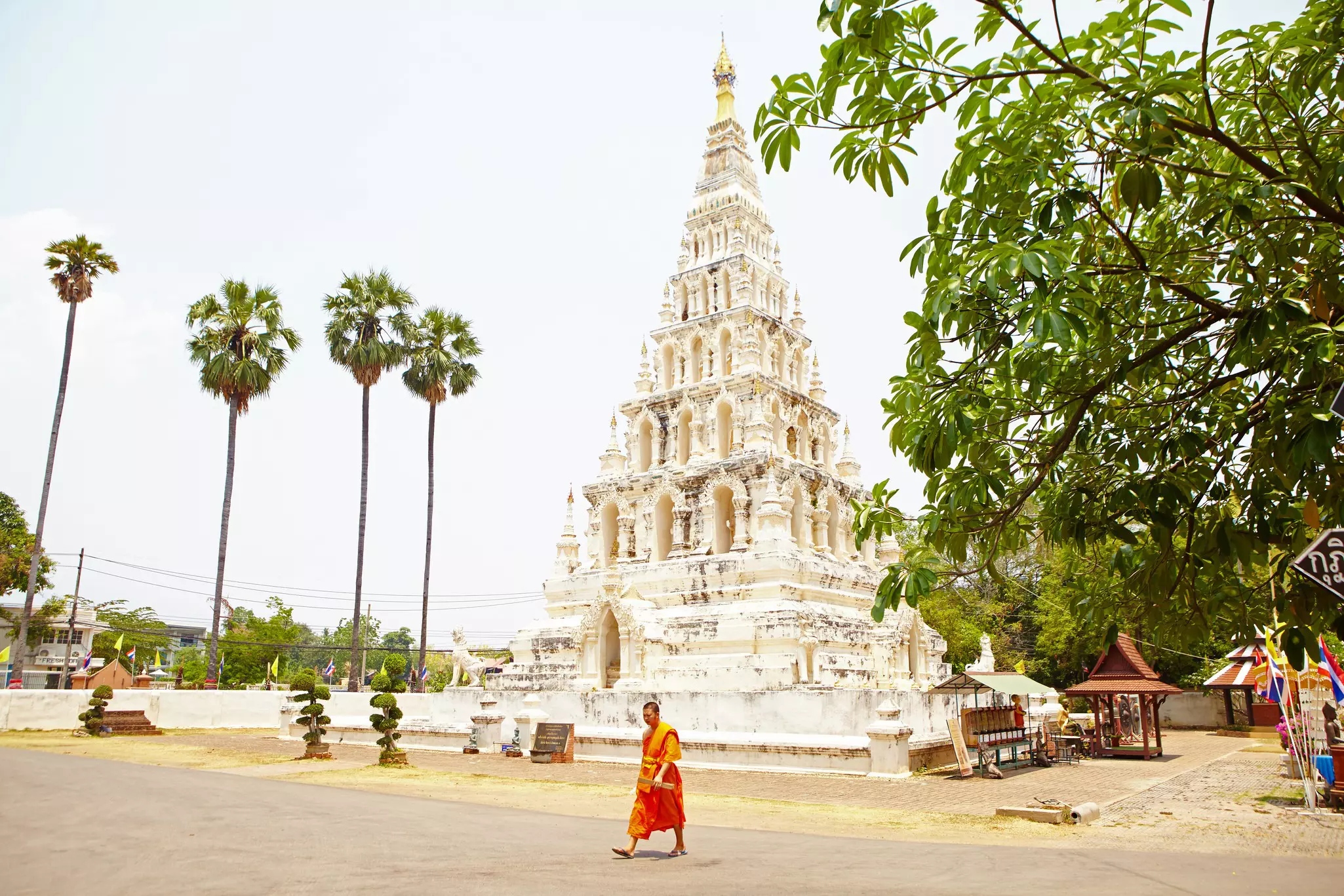 A monk wearing orange robes walk in front of a tall, white, tiered pagoda with a gold top.