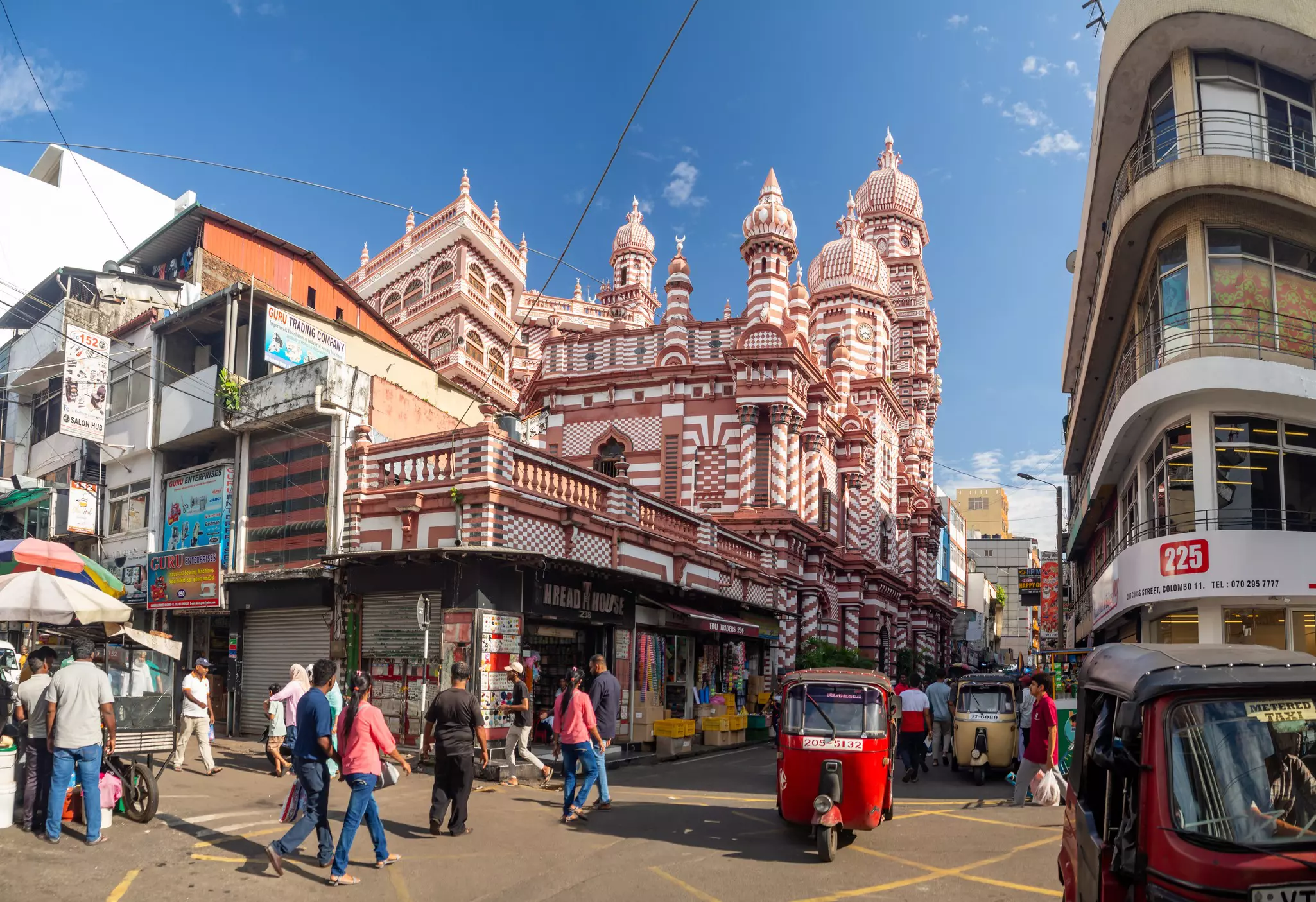 A red tuk tuk passes through a busy neighborhood of shops and markets in front of a distinctive red-and-white striped mosque.