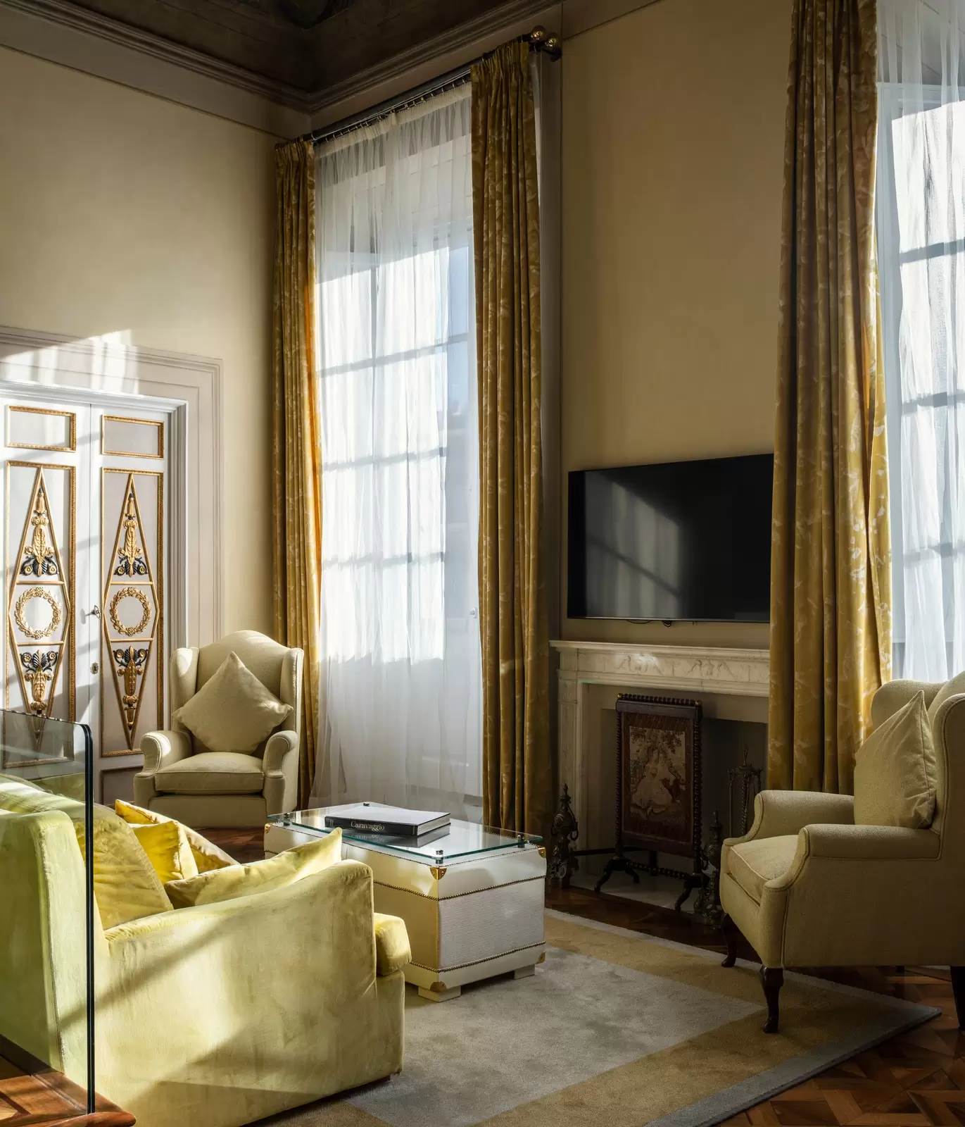 A cozy sitting area with green armchairs and a green couch with a flatscreen TV over a non-operating fireplace, and long curtains over the windows