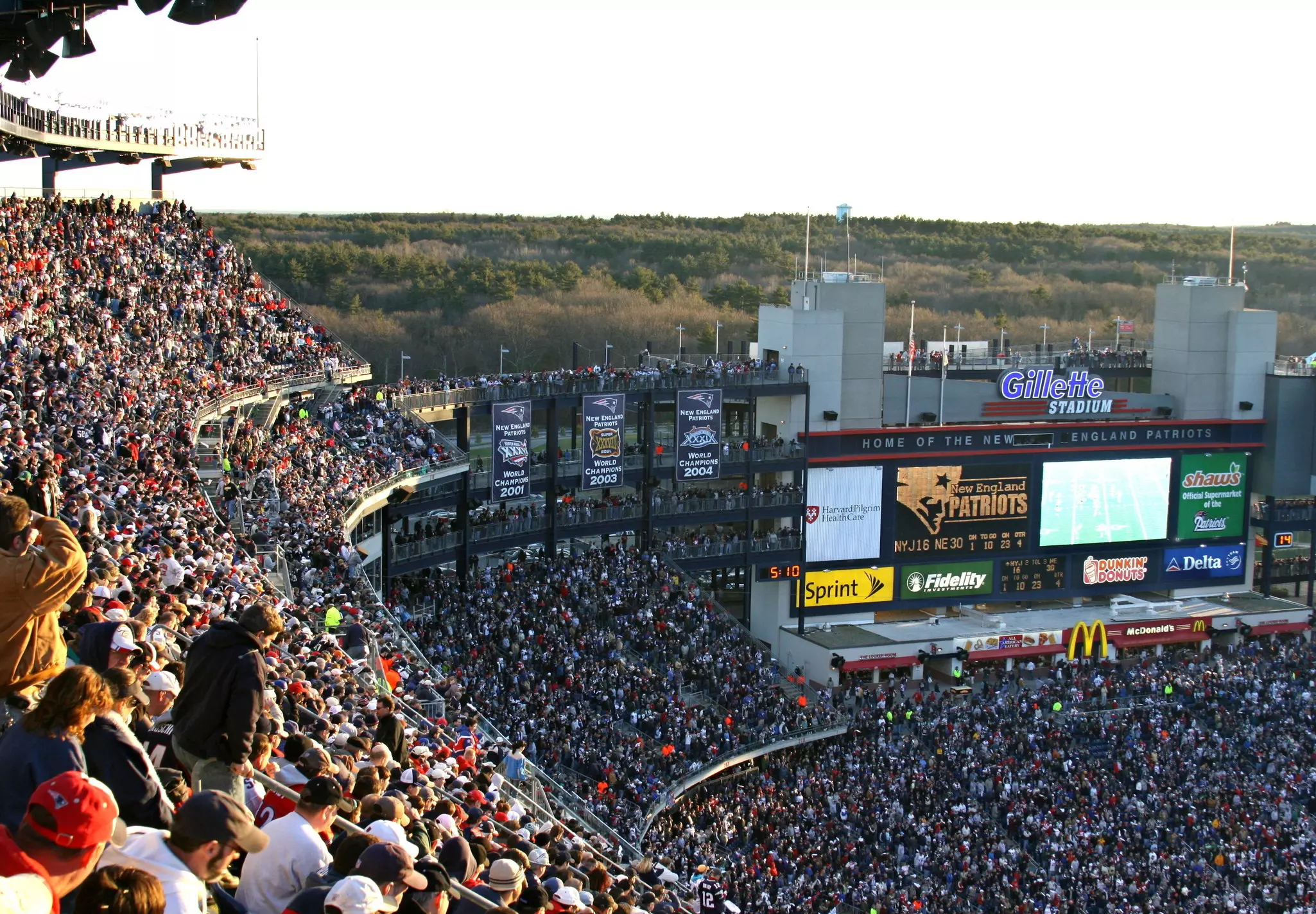 A full football stadium. A giant screen shows action on the field during a New England Patriots game. "Home of the New England Patriots" is across the top of the screen.