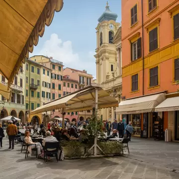 People at café tables under umbrellas in a square of orange and yellow buildings, with a church bell tower behind.