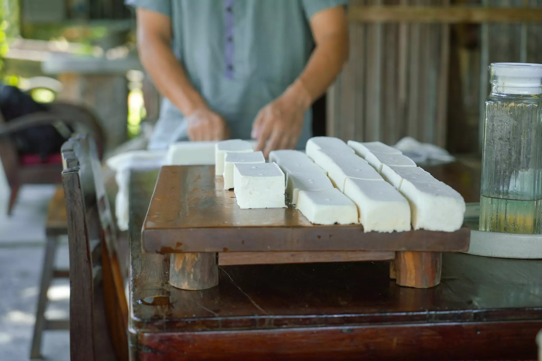 A close-up view of freshly made tofu. A person stands behind the table on which the tofu is displayed.