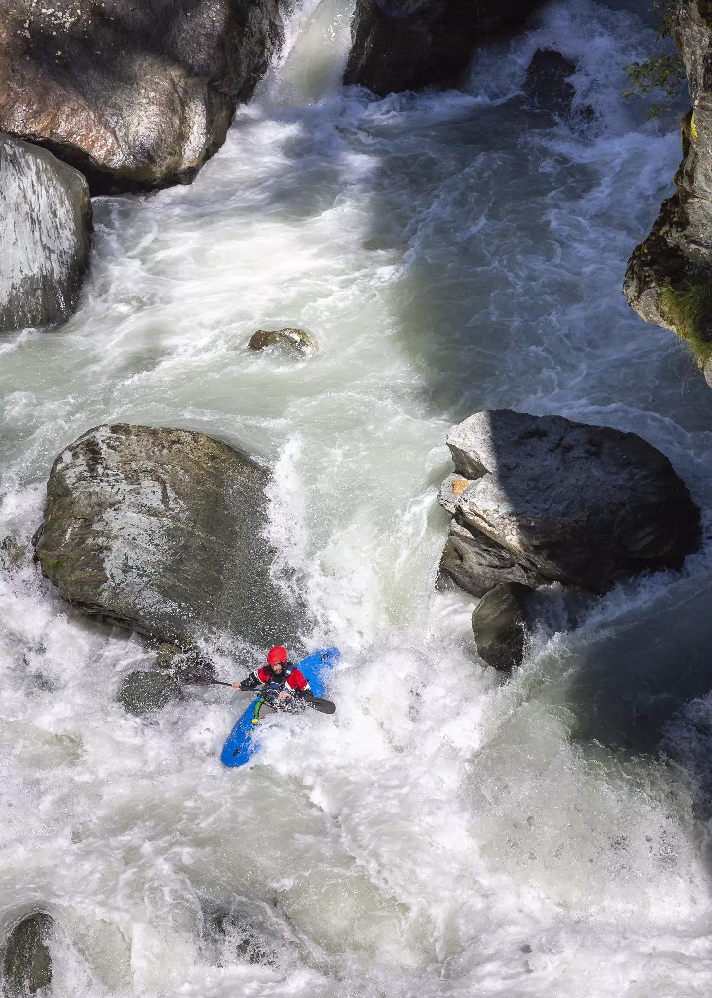 Vertical shot of a male athlete running whitewater rapids in Austria; the water is frothing as it cuts between a series of large boulders in the river.