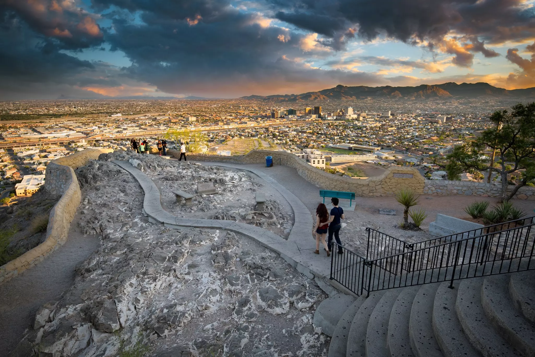 Two people walk toward a panorama in a public park, with a view of a city skyline and mountains beyond seen in dramatic late-day light.