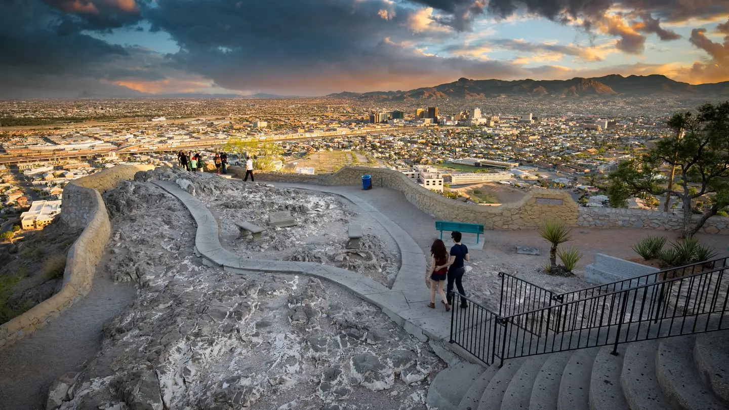 Two people walk toward a panorama in a public park, with a view of a city skyline and mountains beyond seen in dramatic late-day light.