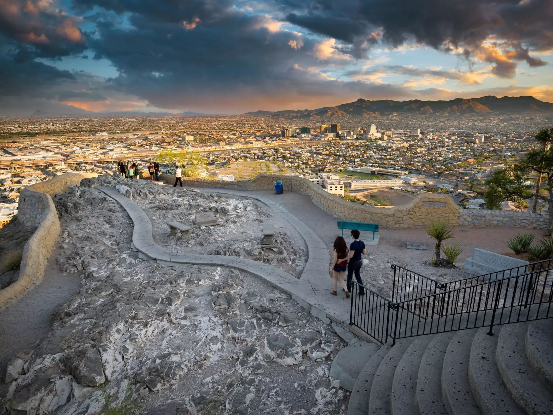 Two people walk toward a panorama in a public park, with a view of a city skyline and mountains beyond seen in dramatic late-day light.