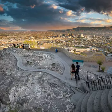 Two people walk toward a panorama in a public park, with a view of a city skyline and mountains beyond seen in dramatic late-day light.