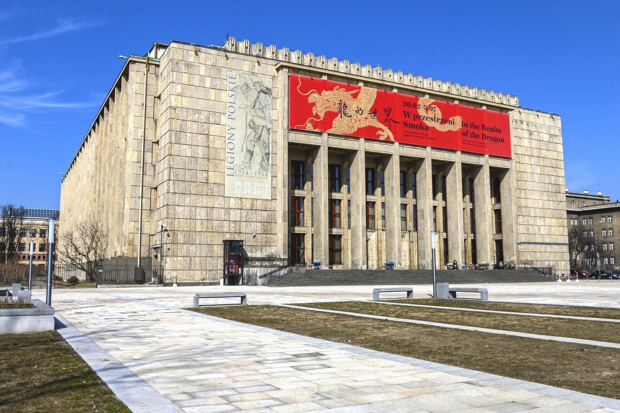 A monolithic 1930s limestone building with a deep pillared portico, hung with a large red exhibition banner, above an empty paved forecourt.
