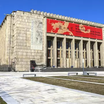 A monolithic 1930s limestone building with a deep pillared portico, hung with a large red exhibition banner, above an empty paved forecourt.