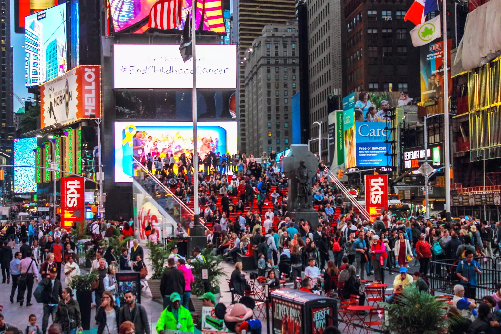 People crowd a busy city square, surrounded by electronic billboards on skyscrapers.