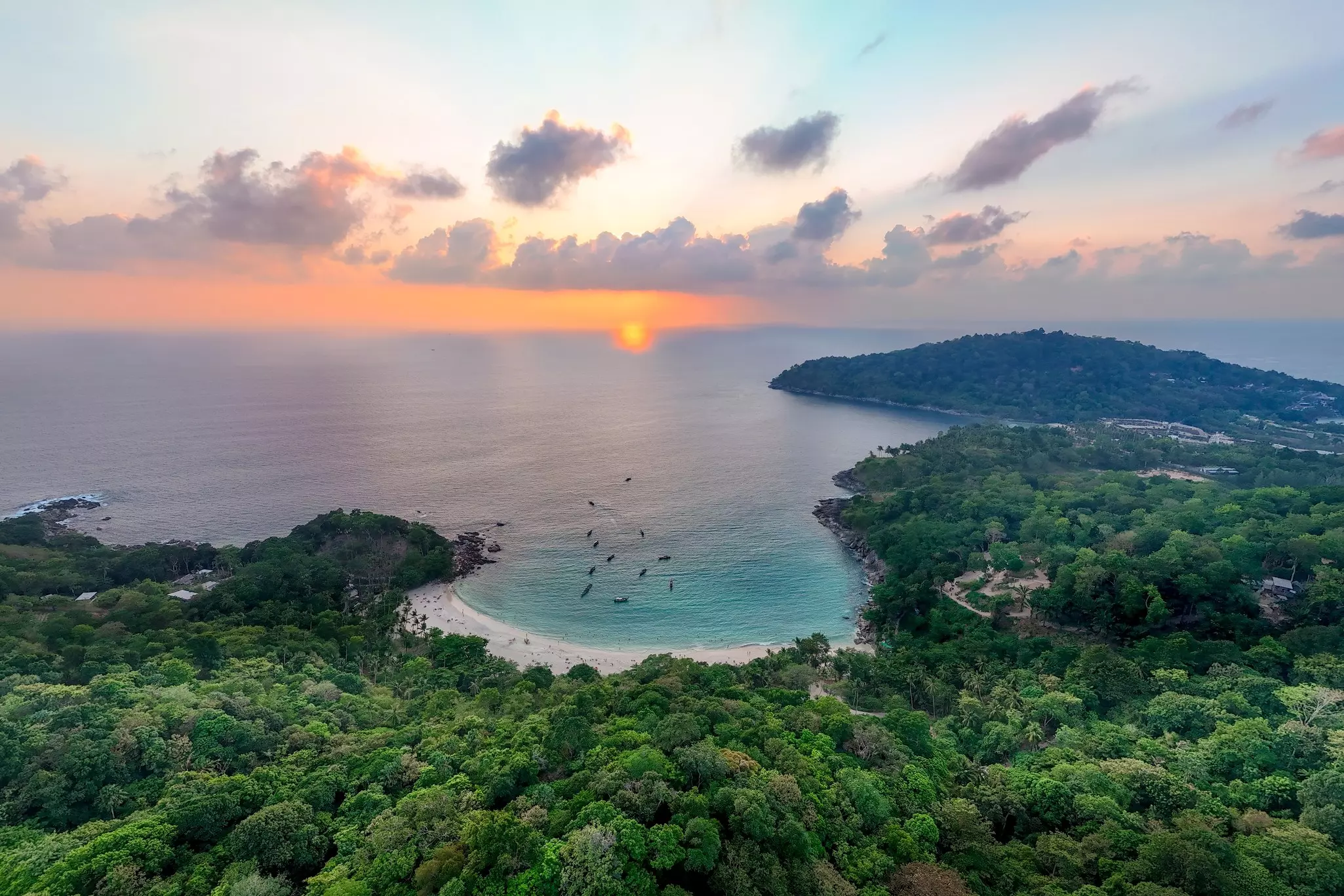 A sheltered bay backed by dense jungle at sunset. Small boats are moored in the ocean 