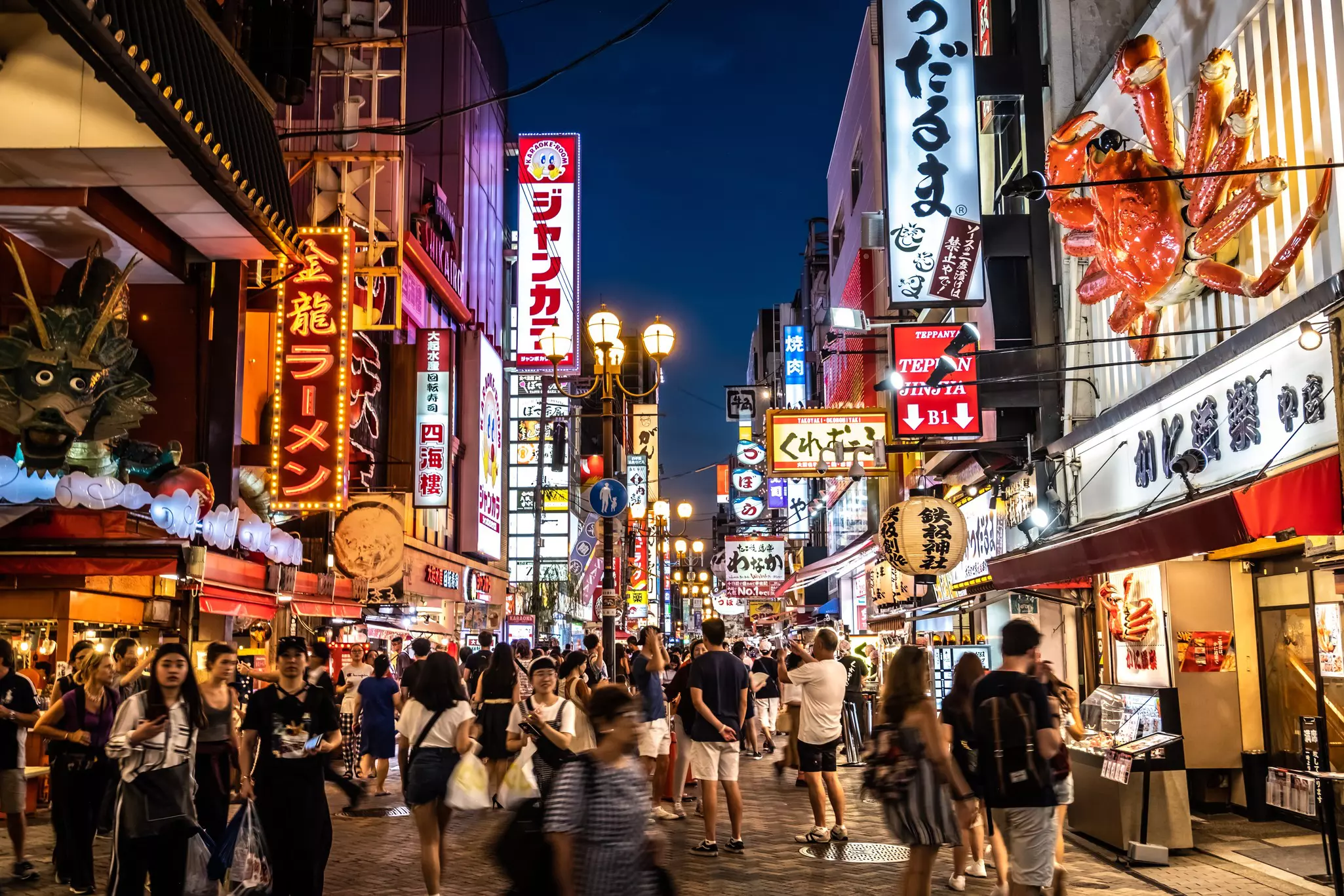 People crowd in a street a night. Numerous neon signs mounted on the buildings illuminate the scene.