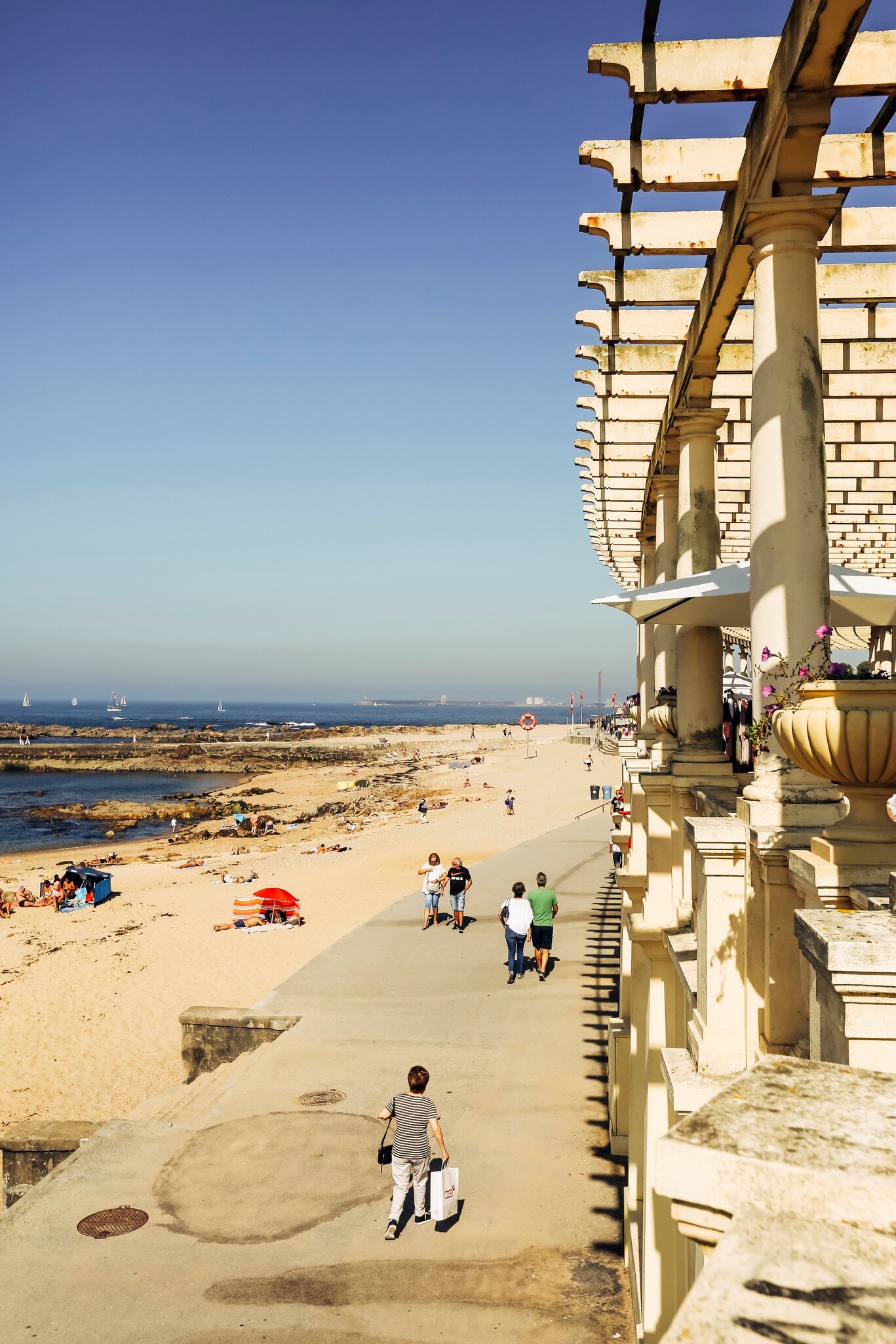 White stone pergola beside a seafront promenade with walkers, a sandy beach and the Atlantic below, Foz do Douro, Porto.
