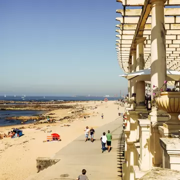 White stone pergola beside a seafront promenade with walkers, a sandy beach and the Atlantic below, Foz do Douro, Porto.