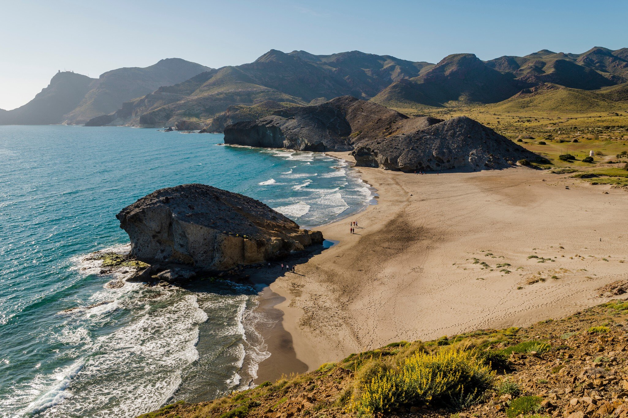 A sandy beach surrounded by cliffs. A freestanding rock marks the centre of the shoreline. 