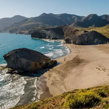 A sandy beach surrounded by cliffs. A freestanding rock marks the centre of the shoreline. 