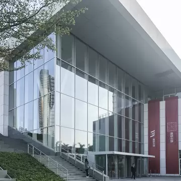 Glass-and-white stone exterior of Design Society in Shenzhen with red entrance banners and a visitor on the steps.