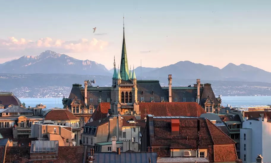 Tiled rooftops and a green-spired stone building in Lausanne, with Lake Geneva and the Alps behind at sunset