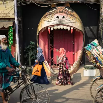 Two women in headscarves pass a giant painted tiger-mouth doorway on a Lalbagh street, Dhaka, cycle rickshaws either side