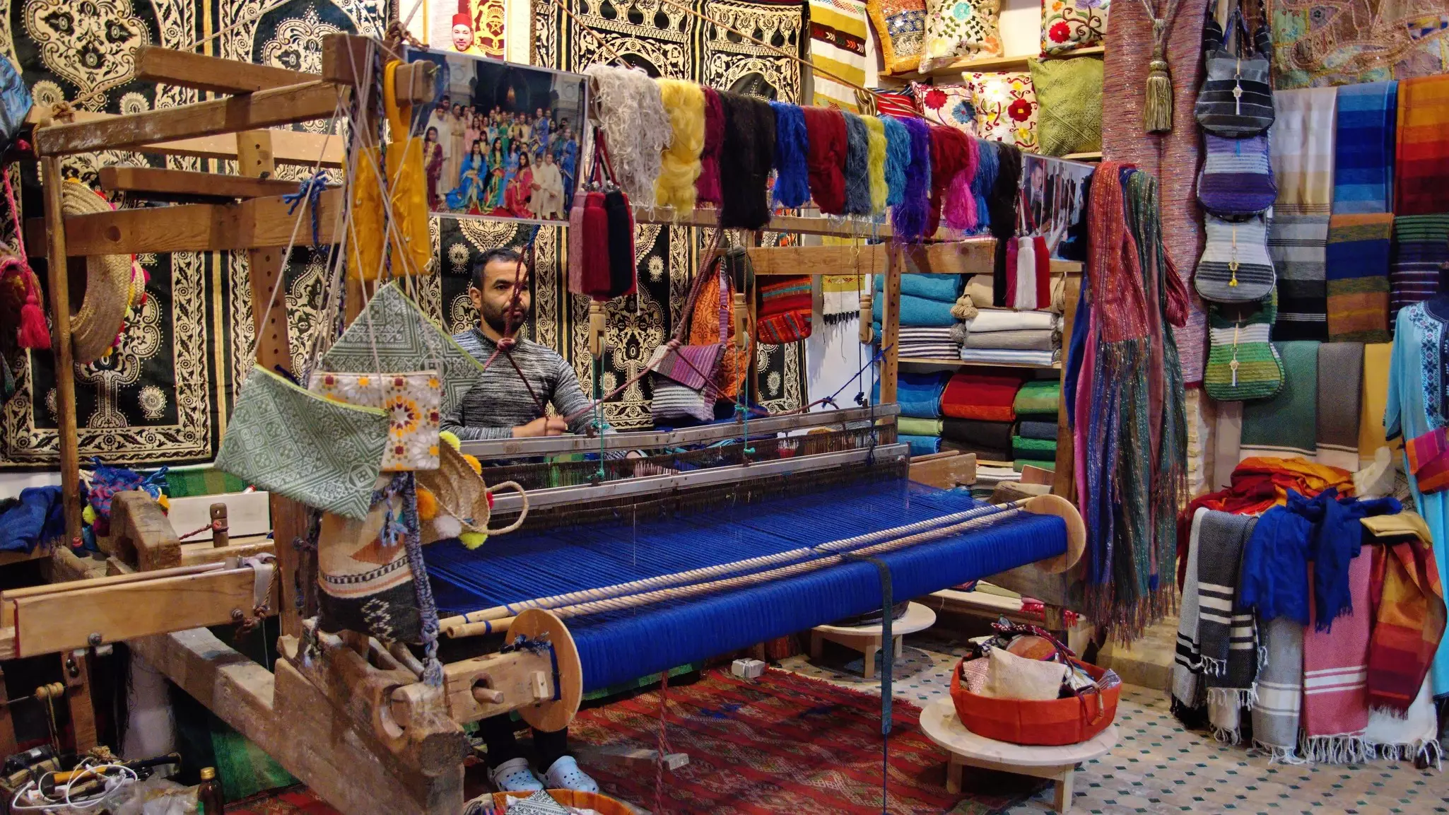 A man sits behind a large loom that is weaving blue thread in a fabric shop.