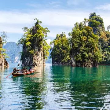 Wooden longtail boat with passengers on green water beside tall limestone rock pillars covered in trees, Khao Sok, Thailand.