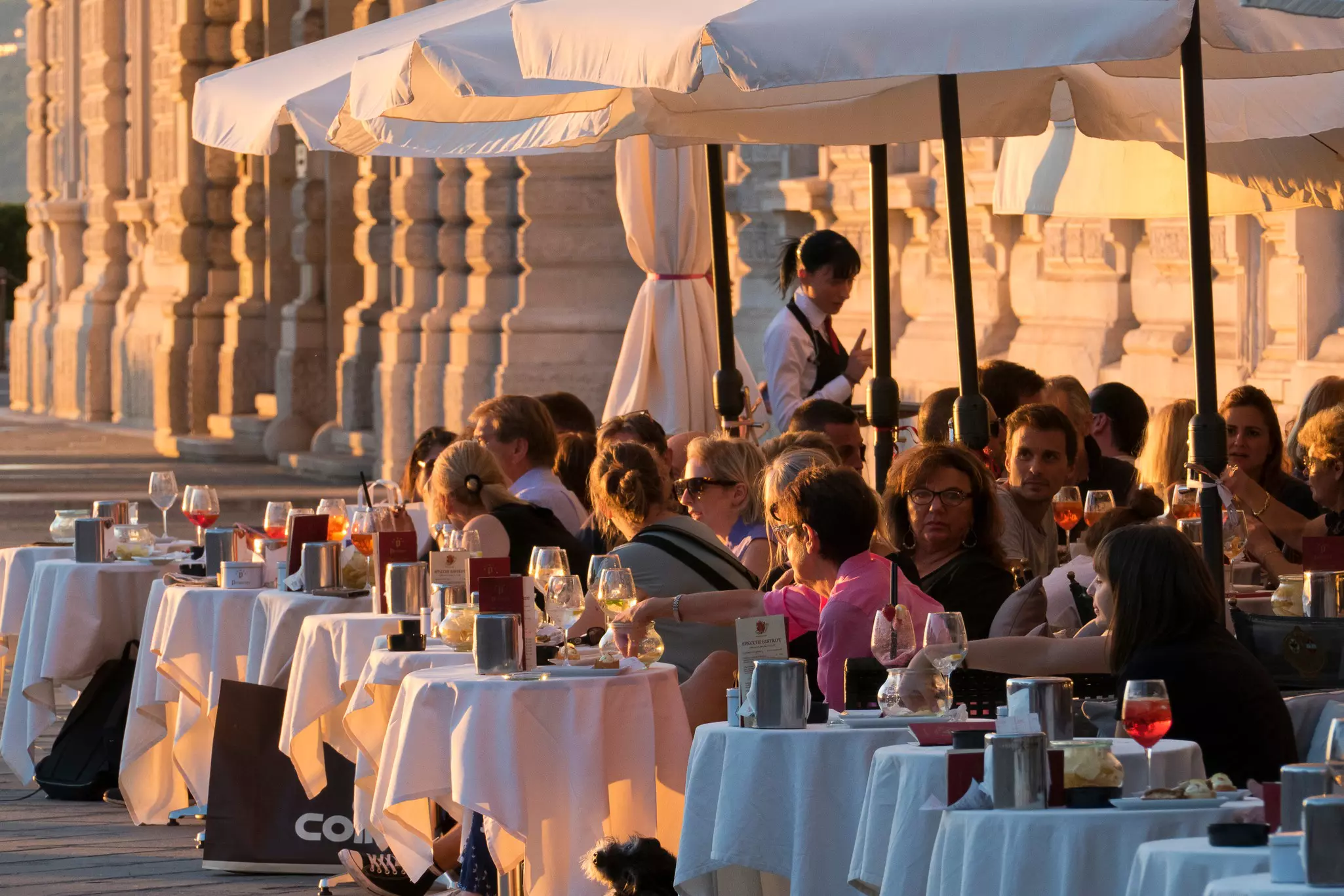 People site at outdoor tables with tablecloths, food and drink. The sunset light illuminates the scene, and the columns behind the tables.