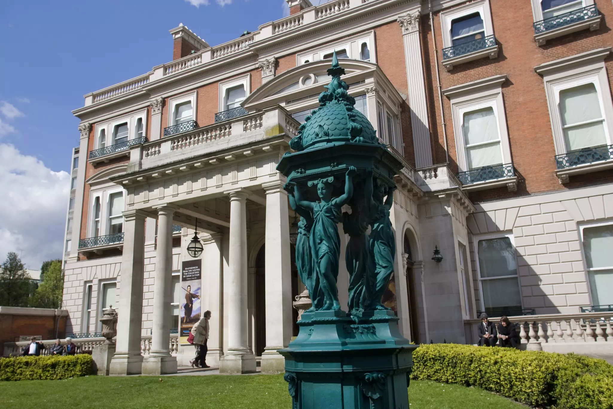 A greenish statue on a lawn in front of the red brick Wallace Collection building in London.