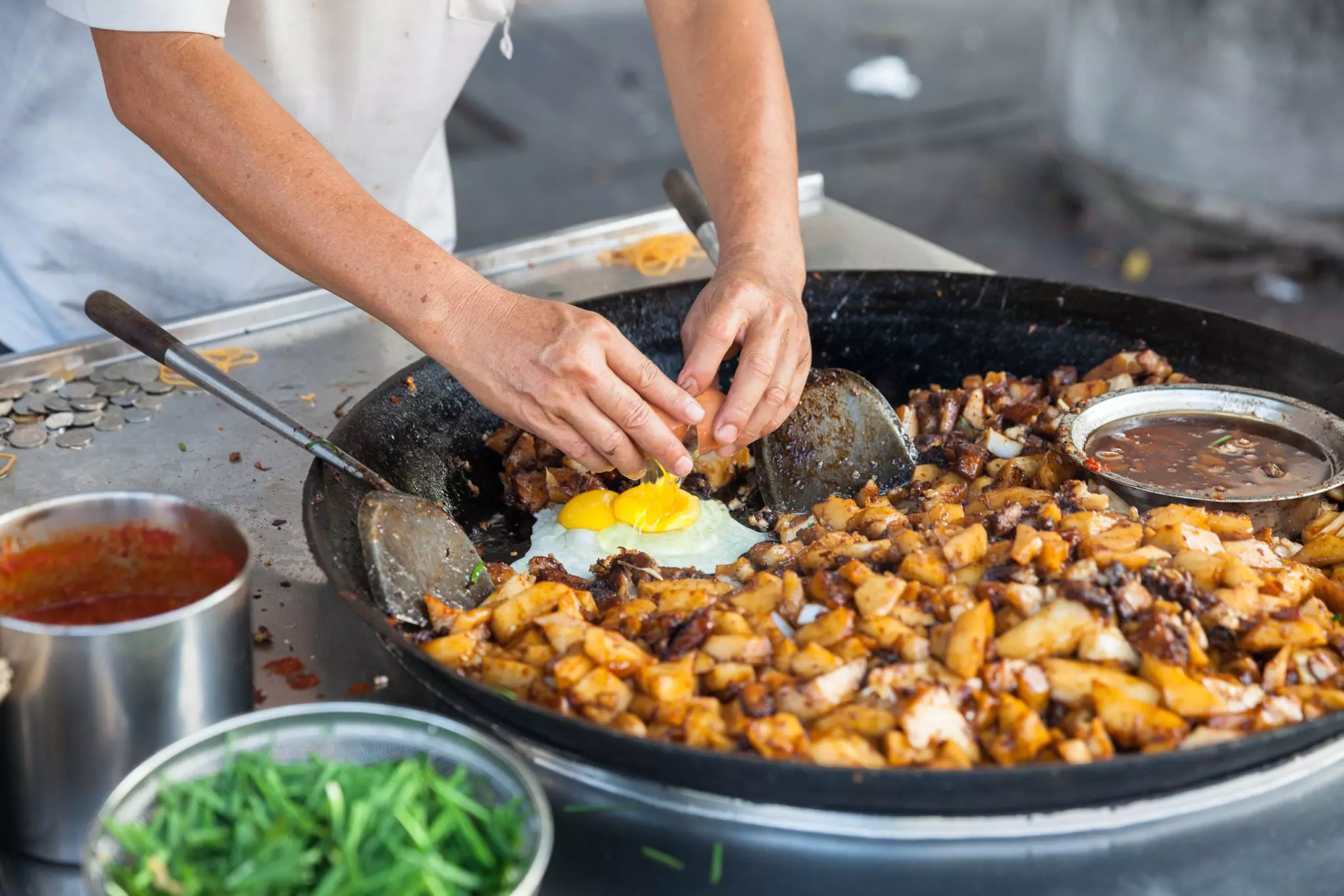 A person breaks an egg at the edge of a large hot plate at a street-food stall.