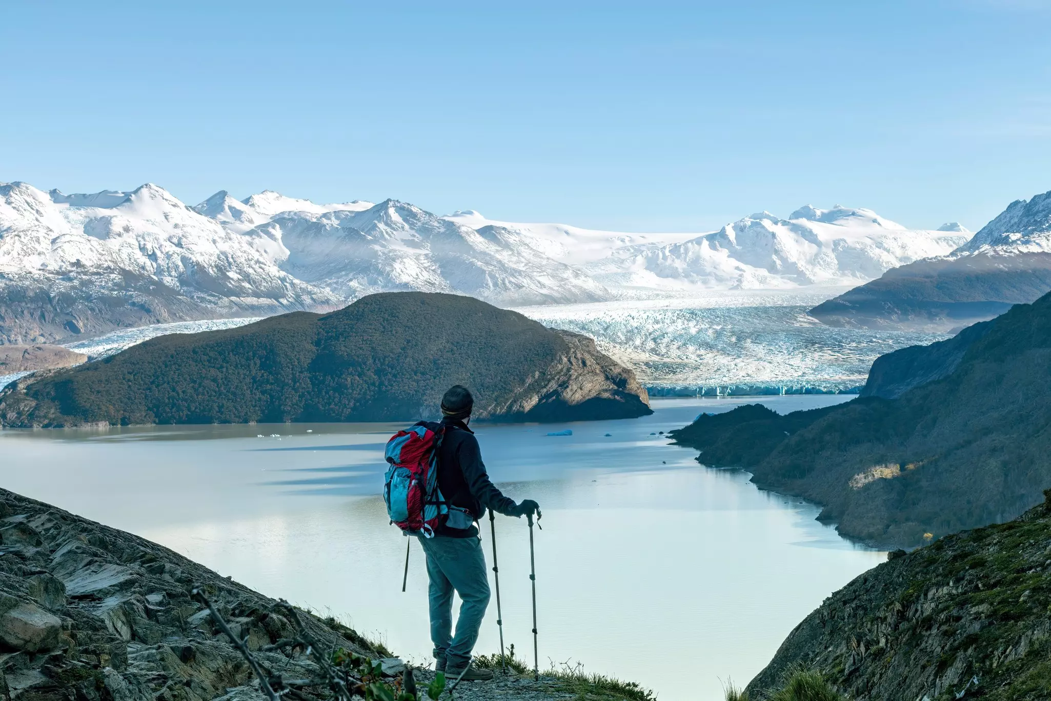 A hiker with poles and a backpacks stands at an overlook with a view of a lake, the face of a glacier and snow-capped mountains in the distance.