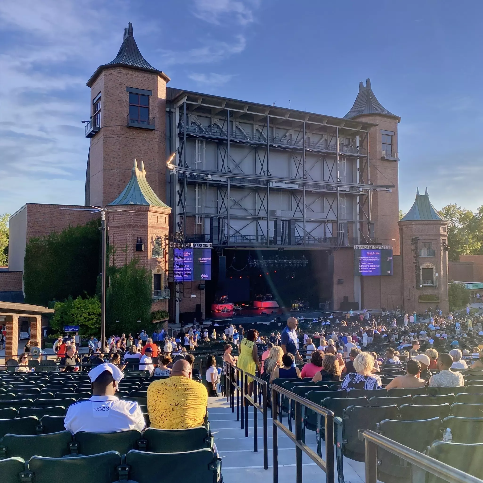 People settle into seats at an outdoor theater before a show. A covered stage with turrets is at the front of the seats.