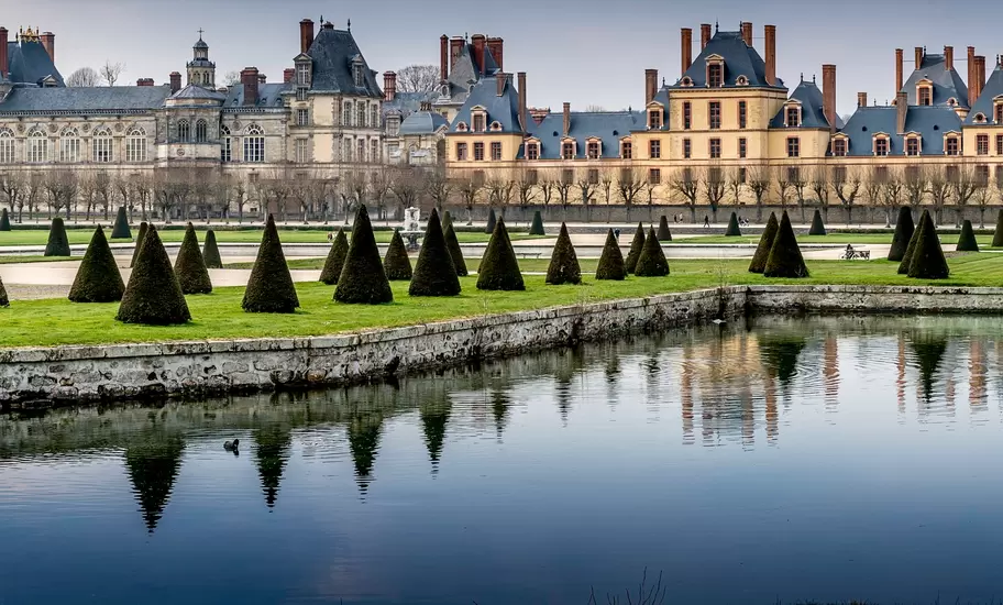 Château de Fontainebleau, France, with cone-shaped topiary on lawns reflected in the still water of the palace pond